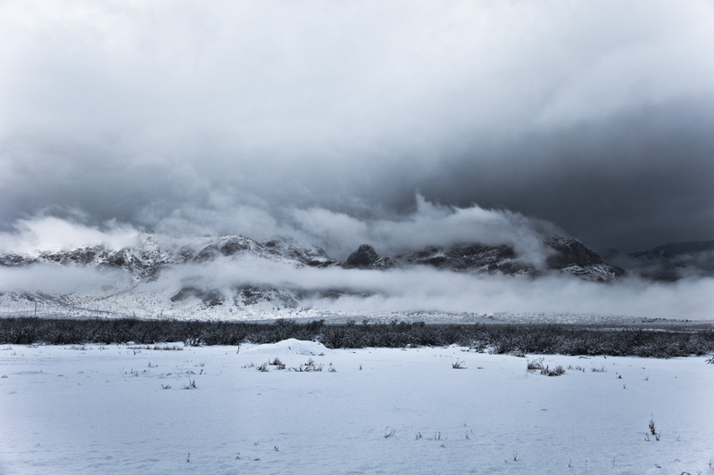 Snow covering the ground and mountains, with dark clouds menacing the sky.