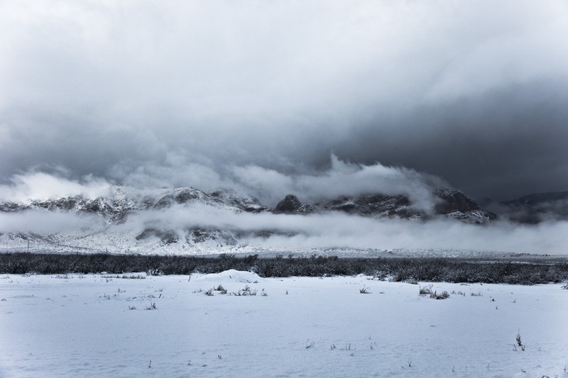 Snow covering the ground and mountains, with dark clouds menacing the sky.