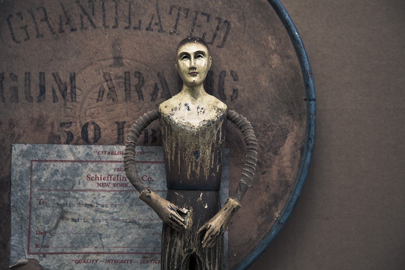 A wooden man standing before the lid to a container for granulated gum arabic.
